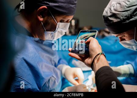 Ritaglio ad angolo elevato maschio che fotografa con smartphone mentre chirurghi concentrati in interventi chirurgici ad esecuzione uniforme utilizzando attrezzi speciali in sala operatoria di ospedale moderno Foto Stock