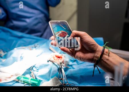 Ritaglio ad angolo elevato maschio che fotografa con smartphone mentre chirurghi concentrati in interventi chirurgici ad esecuzione uniforme utilizzando attrezzi speciali in sala operatoria di ospedale moderno Foto Stock