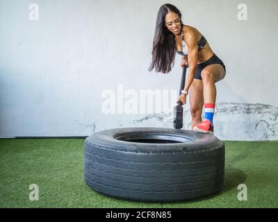 Sicuro allegro forte atleta adulta in sportswear mettendo a fuoco mentre lavorare con un martello pesante e uno pneumatico su un tappeto verde contro la parete bianca nel club sportivo Foto Stock