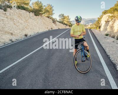 uomo sano che riposa e usa lo smartphone dopo aver cavalcato una bicicletta su una strada di montagna in una giornata di sole Foto Stock