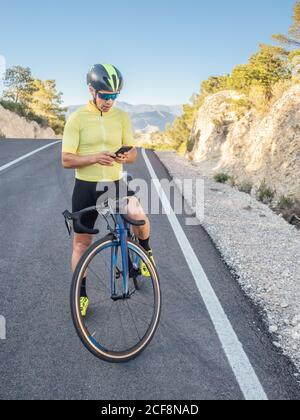 uomo sano che riposa e usa lo smartphone dopo aver cavalcato una bicicletta su una strada di montagna in una giornata di sole Foto Stock