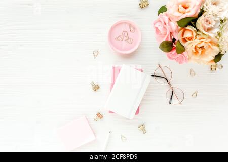 Spazio di lavoro femminile con fiori rosa e beige rose bouquet, accessori, diario, bicchieri su sfondo bianco. Disposizione piatta, vista dall'alto. Concetto di blog di bellezza. Foto Stock