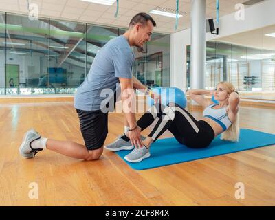 Vista laterale del personal trainer che aiuta la giovane donna a fare scricchioloni addominali sul pavimento in una spaziosa palestra Foto Stock