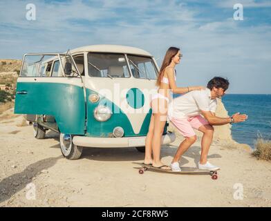 Vista laterale di padre e figlia che giocano a bordo lungo vicino rimorchio colorato sulla spiaggia collinare polverosa sullo sfondo di sereno acqua Foto Stock