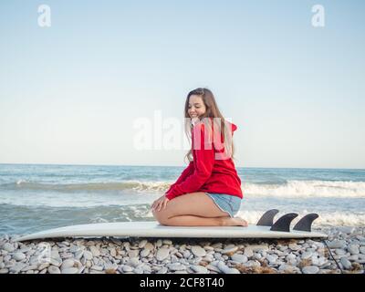 Vista laterale di attraente sorridente donna a capelli lunghi in felpa con cappuccio rosso godendosi seduto sulla tavola da surf sul mare roccioso Foto Stock