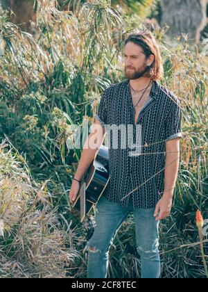 Uomo hipster pusivo che cammina nella giungla con la chitarra che guarda alla telecamera Foto Stock