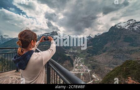 Vista posteriore Donna in abiti caldi in piedi sul molo e guardando cresta di montagna e scattare foto con il telefono cellulare in campagna Foto Stock