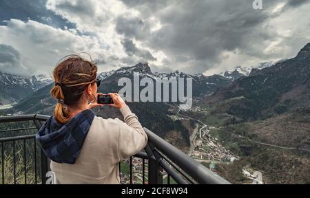 Vista posteriore Donna in abiti caldi in piedi sul molo e guardando cresta di montagna e scattare foto con il telefono cellulare in campagna Foto Stock