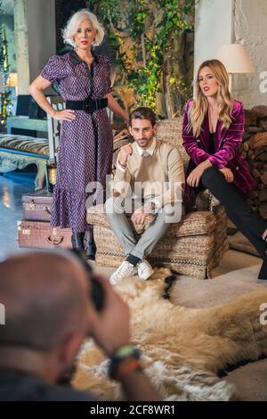 Basso angolo di uomo sfocato senza volto che scatta con macchina fotografica della signora grigia grave con capelli in elegante vestito in piedi accanto ad adulto maschio e femmina seduta contro casa di campagna interno in studio fotografico Foto Stock