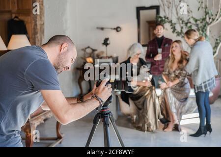 Vista laterale della messa a fuoco e della preparazione della telecamera da parte dell'uomo adulto per scattare mentre si è in piedi contro il lavoro sfocato dell'artista di compongono con modelli in casa di campagna in stile vintage Foto Stock