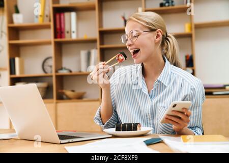 Immagine della donna bionda felice che mangia sushi con i chopsticks mentre utilizzo di cellulare e computer portatile in ufficio Foto Stock