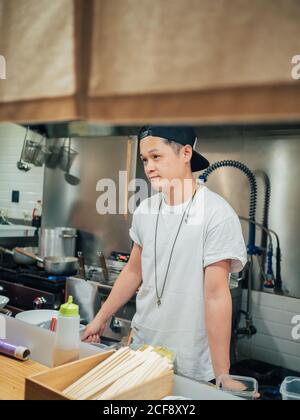 Giovane uomo in maglietta bianca e cappellino nero ramen cucina In ristorante giapponese Foto Stock