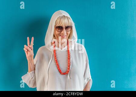 Ritratto di bella donna anziana dai capelli bianchi con occhiali da sole in camicia bianca con cappuccio sulla testa e perle di corallo su sfondo parete blu Foto Stock