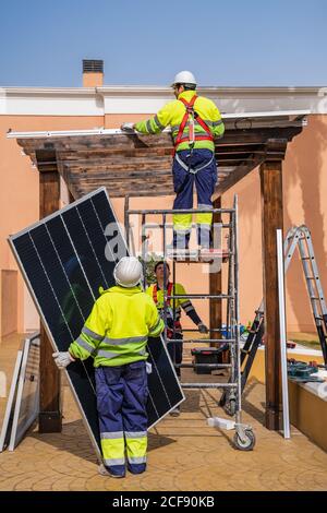 Gruppo di lavoratori in uniforme e caschi che installano pannelli fotovoltaici sul tetto di costruzione in legno vicino a casa Foto Stock