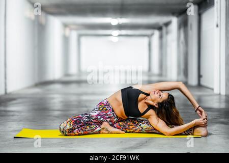 Vista laterale della donna adulta in rivoluzione testa a ginocchio posa mentre si pratica yoga in garage leggero sala guardando verso l'alto Foto Stock