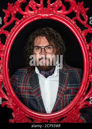 Uomo serio e ben vestito con capelli ricci e barba posa in cornice rossa su sfondo nero Foto Stock