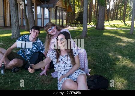 Un gruppo di persone prende un selfie seduto sul prato nel parco, tre amici sorridendo guardando lo smartphone fotocamera Foto Stock