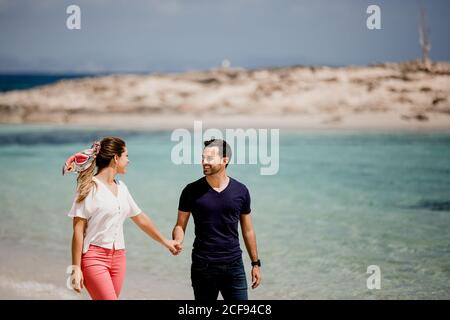 Sorridendo uomo e donna che camminano lungo la riva del mare mentre si tengono le mani e si guardano con amore su sfondo sfocato Foto Stock