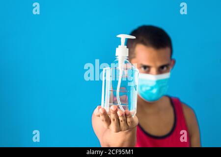 Giovane maschio in una maschera protettiva che tiene una mano igienizzatore isolato su sfondo blu Foto Stock