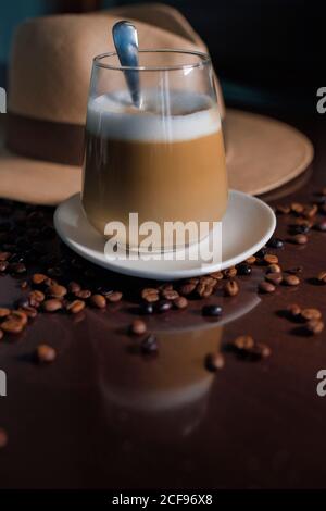 Deliziosa bevanda marrone aromatica con schiuma bianca in vetro sopra piatto rotondo in piedi tra chicchi di caffè arrosto accanto al cappello con brim su tavolo lucido in legno Foto Stock