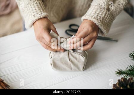 Donna in felpa accogliente preparazione di plastica regalo di natale gratuito, vacanze zero rifiuti. Mani femminili che avvolgono il regalo di natale in tessuto di lino su legno ta Foto Stock