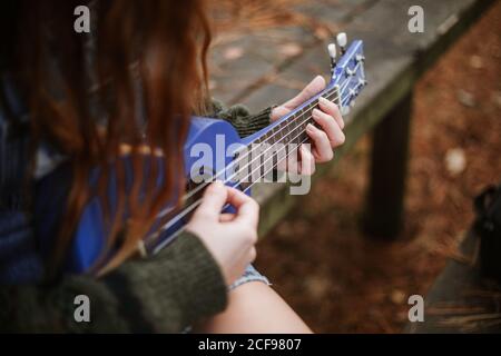 Da sopra brunette irriconoscibile in abiti casual che giocano blu piccolo chitarra mentre si siede da solo su una panchina di legno nella città d'autunno parcheggio Foto Stock