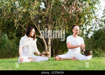 Coppia flessibile e rilassata in stile bianco seduta in posa lotus tenere in mano la ciotola tibetana mentre medita e ti godi tempo insieme nella giornata estiva nel parco Foto Stock