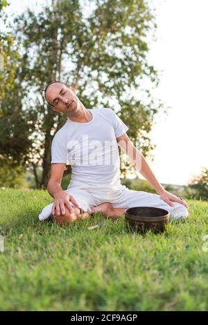 Vista laterale del calmo giovane maschio in bianco indossare seduta Sulle ginocchia e tenendo in mano la ciotola tibetana che canta andare yoga e rilassarsi sul prato in estate Foto Stock