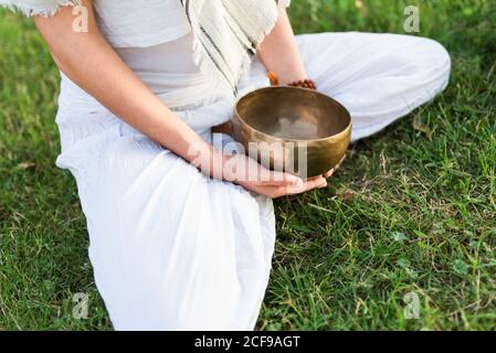 Vista laterale del calmo giovane maschio in bianco indossare seduta Sulle ginocchia e tenendo in mano la ciotola tibetana che canta andare yoga e rilassarsi sul prato in estate Foto Stock