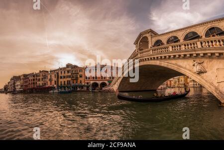 Venezia, Italia - Giugno 10 2019:Barca lunga con una stretta navigazione a vela sotto l'antico ponte lungo il canale d'acqua profonda contro la riva urbana in un giorno nuvoloso Foto Stock