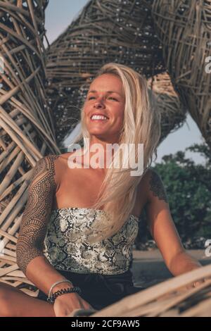 Allegro giovane biondo che indossa abiti estivi casual con tatuaggi le spalle poggiano sui sedili in vimini mentre si guarda al sole giorno d'estate Foto Stock