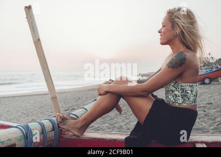 Vista laterale di una giovane donna positiva in un abito casual seduto su una panchina di legno sulla spiaggia di sabbia e ammirando il mare ondulato durante il tramonto in estate Foto Stock