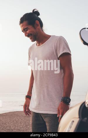 Felice adulto etnico maschio in T-shirt casual in piedi accanto in moto al mare all'ora del tramonto Foto Stock