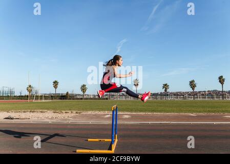 Vista laterale di una giovane donna forte in abbigliamento sportivo che saltella sopra ostacoli contro il cielo blu durante l'allenamento allo stadio Foto Stock