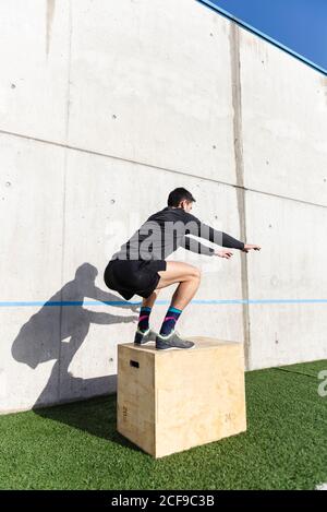 Vista posteriore di giovane atletico maschio che salta sulla scatola vicino parete grigia in giornata di sole Foto Stock