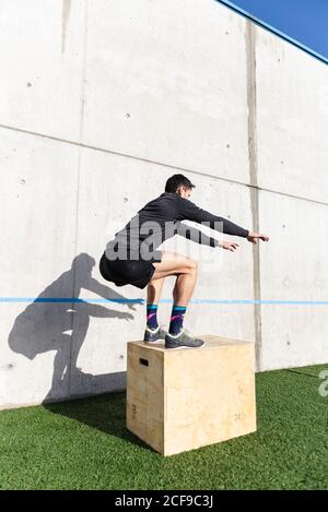 Vista laterale del giovane atletico maschio che salta sulla scatola vicino parete grigia in giornata di sole Foto Stock