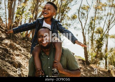 Ragazzo carino seduto sulla spalla di suo padre su un sentiero forestale. Padre e figlio si divertono nelle loro vacanze. Foto Stock