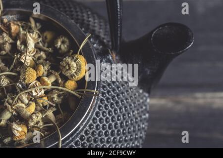 Primo piano di un cumulo di daisy essiccato per la preparazione del tè in metallo teiera su tavolo scuro Foto Stock