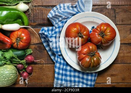 Vista dall'alto del vecchio piatto con grossi pomodori rossi su tovagliolo di tessuto blu e bianco su tavola di legno tavolo con verdure fresche miste messe da parte Foto Stock