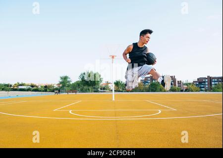 Giovane uomo che gioca sul campo di pallacanestro giallo all'aperto saltando felicemente Foto Stock