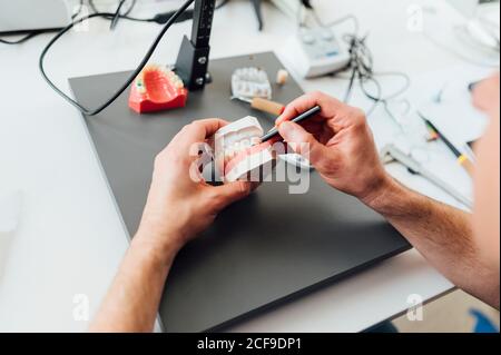 Dall'alto di ortodontista raccolto con matita in lavorazione a mano con stampo dentale bianco a tavola con attrezzatura professionale Foto Stock