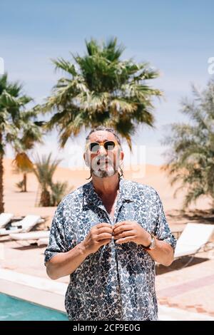 Uomo anziano con bearded in occhiali da sole che bottonano la camicia mentre si sta sopra piscina nella giornata di sole in resort Foto Stock