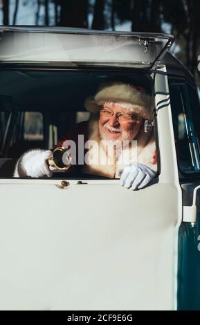 Uomo anziano sorridente in costume di Babbo Natale che guida vecchio furgone con campana in metallo in mano in giornata di sole guardando via Foto Stock