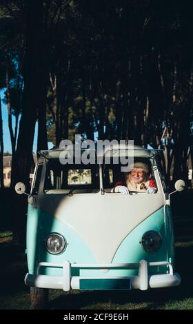 Uomo anziano sorridente in costume di Babbo Natale che guida vecchio furgone con campana in metallo in mano in giornata di sole guardando via Foto Stock