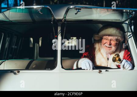 Uomo anziano sorridente in costume di Babbo Natale che guida vecchio furgone con campana in metallo in mano in giornata di sole guardando via Foto Stock