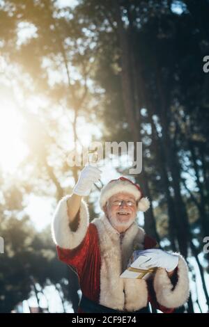 Uomo anziano allegro in costume di Babbo Natale in piedi con presentare e campanello in mani guelate guardando la fotocamera su natura sfondo Foto Stock