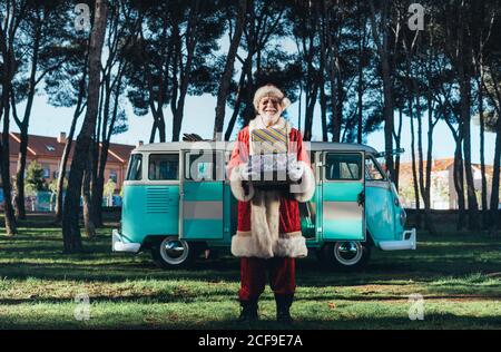 Uomo allegro in costume di Babbo Natale in piedi con regali da furgone turchese su sfondo natura in estate guardando fotocamera Foto Stock
