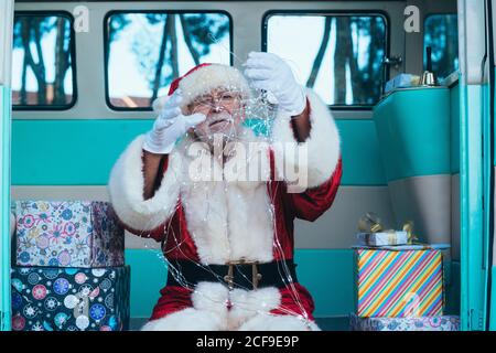 Uomo anziano in costume di Babbo Natale seduto in retrò furgone tra i regali e tenere garland in mani guelse Foto Stock