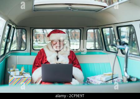 Uomo allegro in costume di Babbo Natale seduto in furgone con computer portatile e regali colorati in giornata di sole Foto Stock