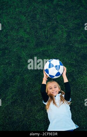 Dall'alto gioiosa presetella in uniforme bianca e blu lanciare palla di calcio mentre si trova da solo su campo verde dentro moderno club sportivo Foto Stock